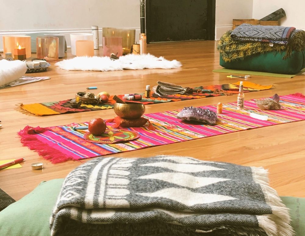 Blanket sitting on bolster pillow in the foreground. Large piece of amethyst, bowls, and other items on two brightly colored, striped mats in the mid ground. White, fuzzy rug and crystal bowls in the background sitting in front of a light colored wall. This is all sitting on a wood floor.